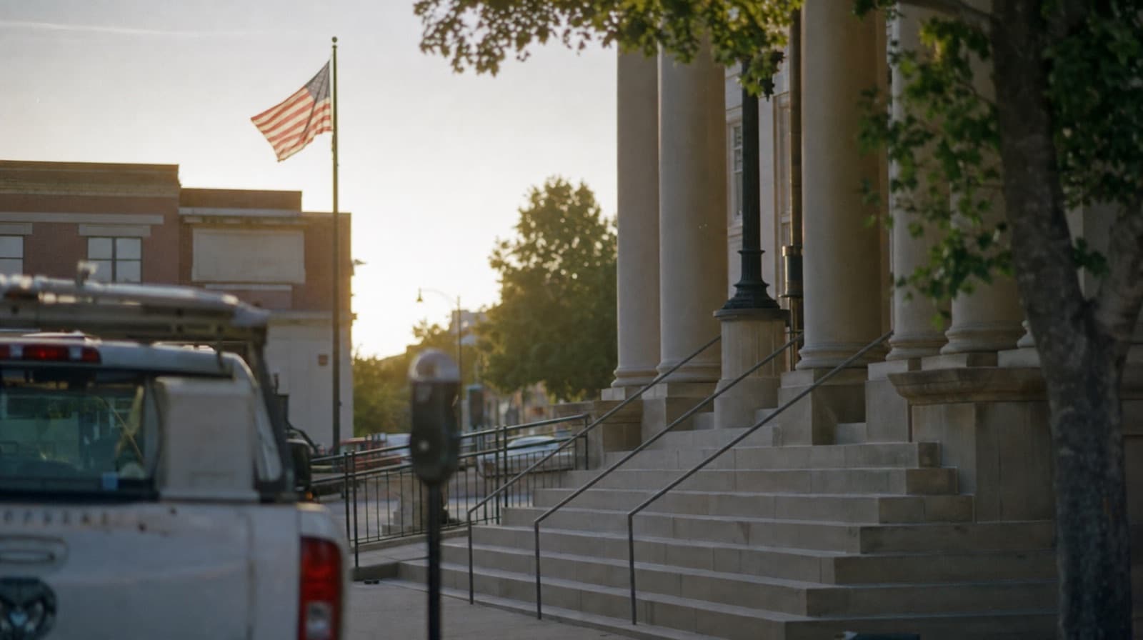 Stone columns and wide stone steps of a civic building, with a brick building across the street and a work pickup parked at the curb in evening light
