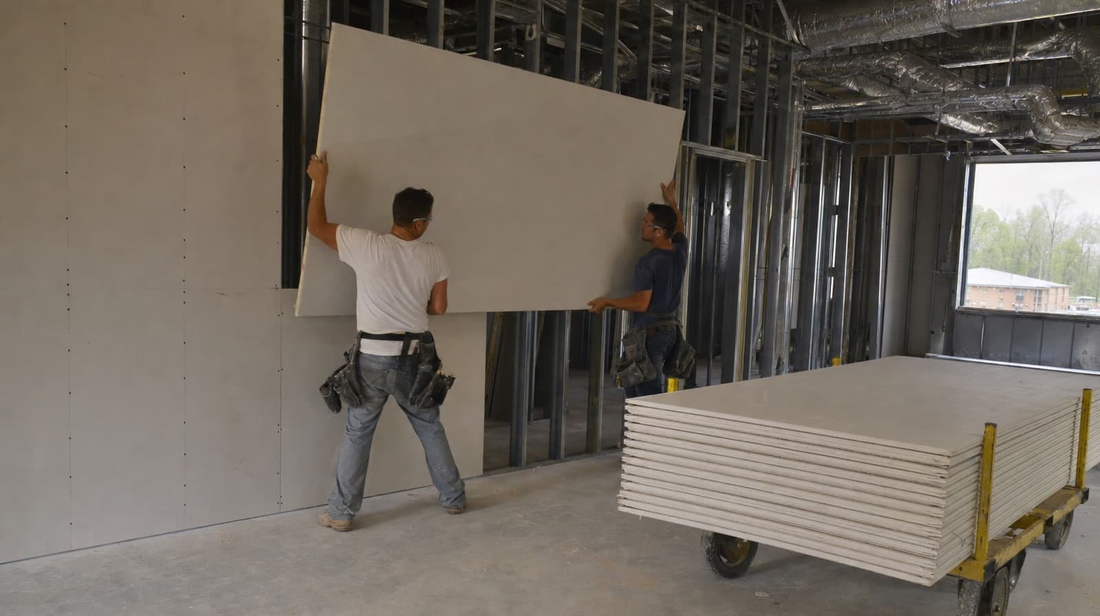 Two drywall installers lifting a gypsum board onto a metal-stud wall beside a cart of stacked drywall sheets