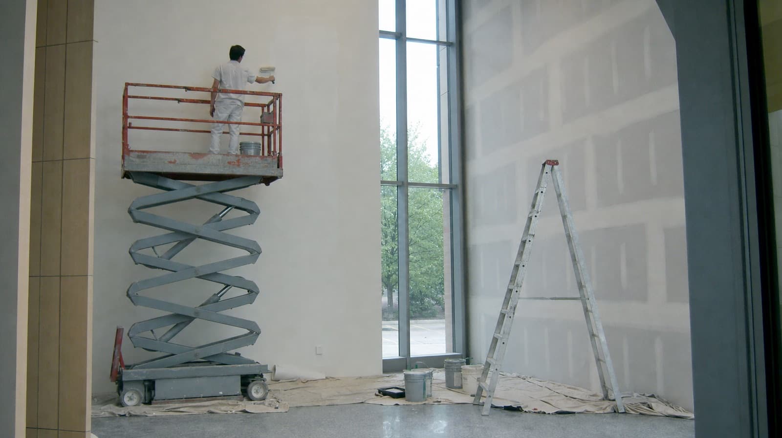 Painter on a scissor lift working on a tall white wall in a new building lobby, with a ladder and drop cloths below